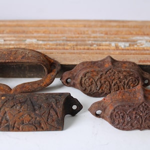 May include: Four antique, rusty metal drawer pulls and a handle. The drawer pulls have ornate floral designs. The handle is a simple oval shape. The items are set against a weathered wooden background.