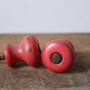 Puede incluir: Dos pomos de madera rojos vintage con tornillos de metal oxidados. Los pomos son redondos con una forma ligeramente bulbosa y un agujero en el centro. Est&aacute;n sobre una superficie de madera envejecida.