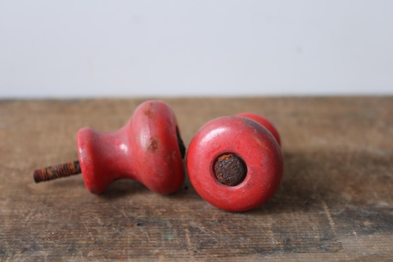Puede incluir: Dos pomos de madera rojos vintage con tornillos de metal oxidados. Los pomos son redondos con una forma ligeramente bulbosa y un agujero en el centro. Est&aacute;n sobre una superficie de madera envejecida.