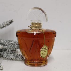 May include: A vintage amber glass perfume bottle with a faceted base and a clear semi-circular stopper. A gold-toned metal collar secures the top. A small, cream-colored paper tag hangs from the neck, printed with Charbert Eau de Toilette New York.