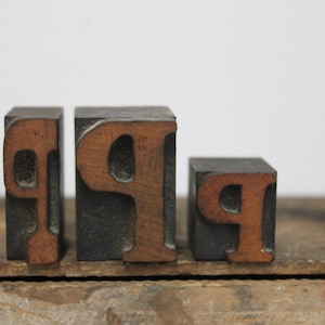 May include: Three vintage letterpress printing blocks made of dark wood and metal, each featuring the lowercase letter p in a different size and font style, arranged in a row with the wooden character faces clearly visible against the dark blocks.