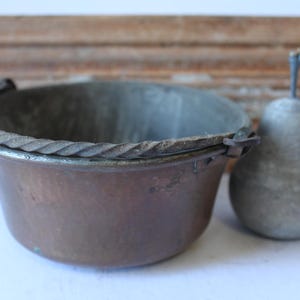 May include: A copper-toned metal bowl with a twisted rope design around the rim, next to a pear-shaped metal object. The bowl has a hammered texture. The background features a wooden plank.