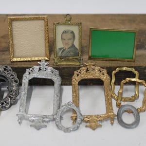 May include: A collection of miniature picture frames in various shapes and metallic finishes, including gold, silver, and dark grey tones. Some frames are empty, one contains a black and white portrait, and another features a solid green insert.