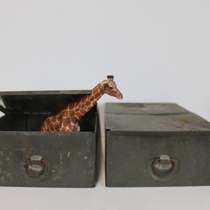 May include: Two weathered, gray metal boxes, one open with a giraffe figurine peeking out. The giraffe has a brown and tan patterned coat. The boxes have ring pulls and are set against a white background.