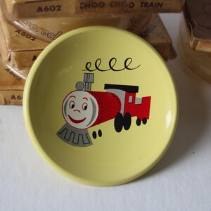 May include: A yellow, circular dish with a cartoon train illustration. The train is red and white with a smiling face. The dish is surrounded by vintage packaging.