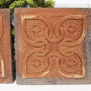 May include: Two square terracotta tiles featuring a raised relief pattern of interlocking curved lines and circular motifs with serrated edges. The tiles have a weathered, matte finish in a reddish-brown hue with visible texture and depth.