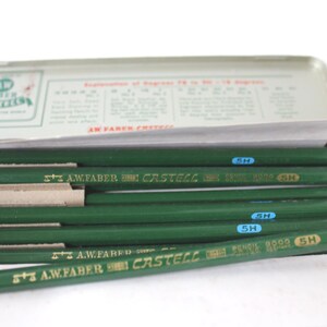 Pode incluir: Uma caixa de l&aacute;pis de metal vintage com tampa articulada, contendo v&aacute;rios l&aacute;pis verdes. Os l&aacute;pis s&atilde;o marcados com "A.W. Faber Castell" e "5H". A caixa tem texto e um logotipo na tampa.