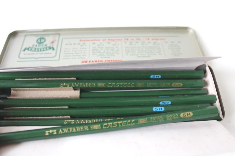 Pode incluir: Uma caixa de l&aacute;pis de metal vintage com tampa articulada, contendo v&aacute;rios l&aacute;pis verdes. Os l&aacute;pis s&atilde;o marcados com "A.W. Faber Castell" e "5H". A caixa tem texto e um logotipo na tampa.