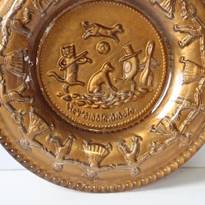 May include: A vintage amber-colored plate with a detailed embossed design. The center features a scene with animal figures and the text "Hey Diddle Diddle". The rim has a repeating pattern of figures.
