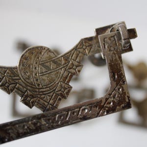 May include: Close-up of antique silver-toned drawer pulls. The pulls have a rectangular frame with decorative geometric and circular patterns. The metal shows signs of wear, adding to their vintage appeal. The background is blurred, highlighting the details of the pulls.
