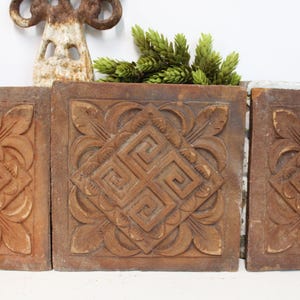 May include: Three square terracotta tiles featuring a raised relief pattern of a central Greek key motif surrounded by stylized leaf designs. The tiles have a weathered, earthy brown finish with visible texture and depth in the carvings.