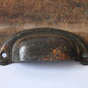 May include: A weathered, antique-style metal drawer pull with a dark, textured finish. The pull has a curved, cup-like design with two mounting holes. The metal shows signs of age with hints of orange rust.