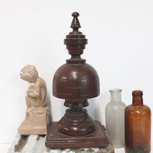 May include: A dark brown wooden finial with a square base and a dome-shaped center featuring carved geometric patterns. Beside it sits a small beige figurine of a seated figure, a clear glass bottle, and a brown glass bottle with a narrow neck.