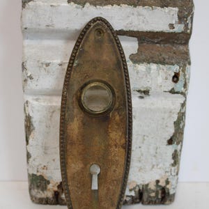 May include: Antique brass oval door plate with a keyhole and a small circular glass window. The door plate is mounted on a weathered, distressed white and gray wooden surface.