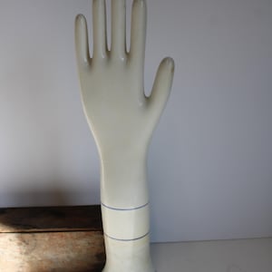 May include: Off-white ceramic glove form with a blue stripe detail around the wrist. The glove form is standing upright, with the fingers extended. The base of the form has the number "805-LB" printed on it.
