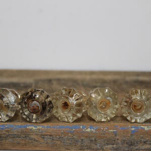 May include: A row of vintage clear glass door knobs with a faceted design. The knobs are arranged on a weathered wooden surface with blue paint. The knobs have a tarnished appearance.