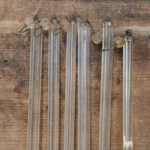 May include: A collection of six clear, vertical glass chandelier prisms, each featuring a faceted rectangular shape. The prisms are suspended by small, dark metal wire loops attached to clear, bead-like glass connectors at the top of each piece.