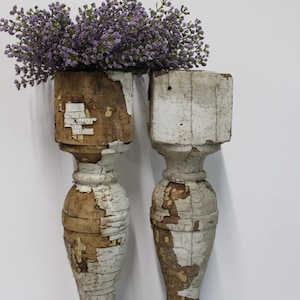 May include: Two tall, weathered wooden architectural balusters with remnants of white paint revealing natural brown wood underneath. The tops of the balusters are flat and square, supporting a cluster of small, purple artificial flowers.