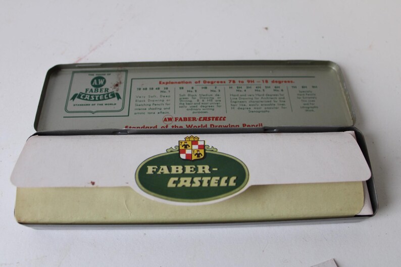 Pode incluir: Um estojo de l&aacute;pis Faber-Castell vintage aberto. A caixa de metal apresenta um logotipo verde e branco e o texto "AW FABER-CASTELL" e "Standard of the World Drawing Pencil". O estojo cont&eacute;m l&aacute;pis.