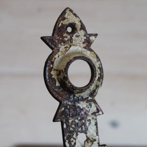 May include: Close-up of a decorative, antique metal piece with a distressed, aged appearance. The metal is primarily brown with areas of peeling white paint, revealing the underlying metal. The design includes a circular element and pointed shapes.