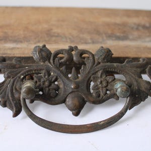 May include: Ornate antique bronze drawer pull with a curved handle and decorative floral and leaf design. The hardware is aged with a patina finish, suggesting vintage or reclaimed origin. The background includes a wooden surface.