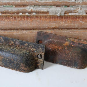 May include: Two antique, weathered metal drawer pulls with a dark, rusty finish. Each pull has a curved handle and two mounting holes. The background features a weathered wooden surface.