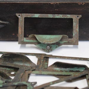 May include: Close-up of antique metal drawer pulls and label holders with a distressed, aged appearance. The metal has a green patina, and the hardware is set against a dark wooden background. These vintage pieces are likely from a cabinet or desk.