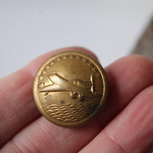 May include: A vintage brass button with an embossed airplane design. The button features a detailed depiction of a plane flying over water, surrounded by stars. The button is round and has a slightly aged appearance.
