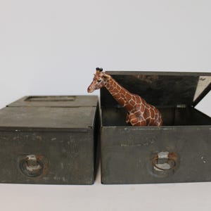 May include: Two olive-green metal storage boxes, one open with a giraffe figurine inside. The giraffe has a brown and tan patterned coat. The boxes have handles and are set against a white background.