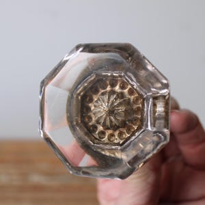 May include: Clear, faceted glass doorknob with a decorative, starburst-like design in the center. The knob has an octagonal shape and is held against a neutral background. This vintage-style knob is suitable for home decor.