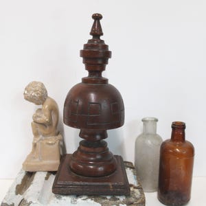 May include: A dark brown wooden finial with a square base and a carved geometric pattern on its dome, accompanied by a small beige figurine of a seated figure, a clear glass bottle, and a brown glass bottle, all arranged together.