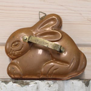 May include: A copper-colored bunny-shaped mold with a hinged brass handle. The mold has a detailed design of a sitting rabbit with long ears and a smiling face. The mold is likely used for baking or making decorative items.