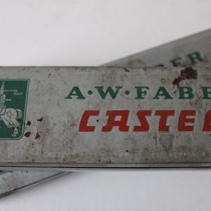 Pode incluir: Duas caixas de l&aacute;pis A.W. Faber Castell vintage. As caixas retangulares de metal s&atilde;o prateadas com letras verdes e vermelhas. Uma caixa tem um bras&atilde;o verde com um cavaleiro a cavalo. As caixas mostram sinais de idade.