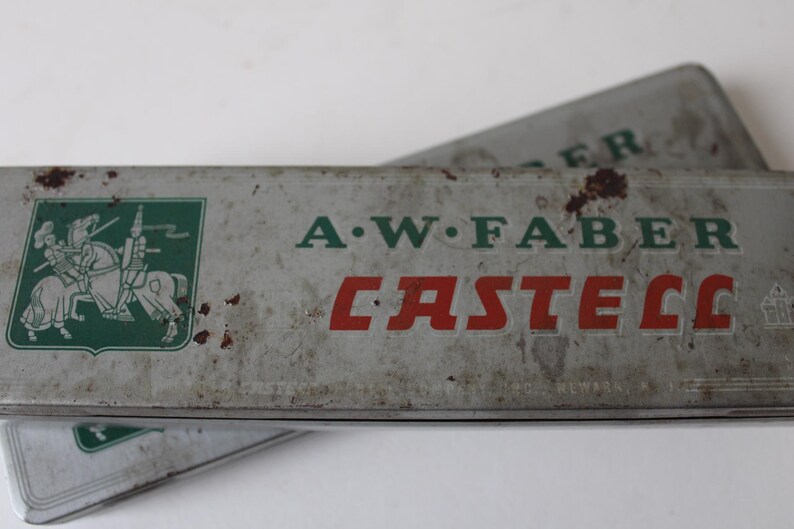 Pode incluir: Duas caixas de l&aacute;pis A.W. Faber Castell vintage. As caixas retangulares de metal s&atilde;o prateadas com letras verdes e vermelhas. Uma caixa tem um bras&atilde;o verde com um cavaleiro a cavalo. As caixas mostram sinais de idade.