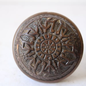 May include: Two antique bronze door knobs with intricate floral and geometric patterns. The knobs are round with a dark, aged finish, showcasing detailed relief designs. The image highlights the craftsmanship of these vintage hardware pieces.