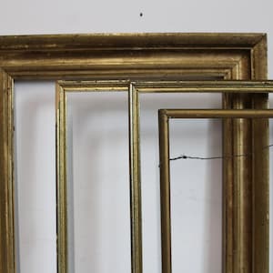 May include: A collection of three rectangular picture frames with a metallic gold finish. The frames vary in width and thickness, featuring worn edges and visible texture. One frame includes a thin metal wire attached to the back for hanging.