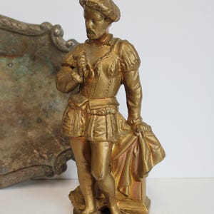 May include: A gold-colored statue of a man in historical attire, including a beret, doublet, and pleated skirt. The figure is holding a cloth and stands on a small pedestal. A decorative, tarnished metal piece is in the background.