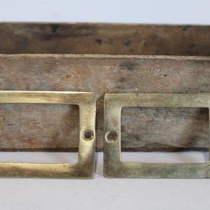 May include: Two rectangular brass label holders with screw holes are displayed against a weathered wooden background. The holders have a vintage, aged appearance, suggesting they are antique or reclaimed. The brass has a warm, golden hue.