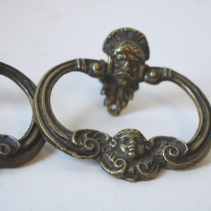 May include: Antique bronze-toned drawer pulls with a classical design. Each pull features a ring handle with a decorative face at the center and a detailed face at the mounting plate. The pulls are likely made of metal and have an aged appearance.