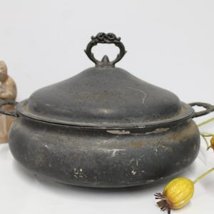 May include: A dark gray, weathered metal tureen with a matching lid featuring a decorative handle. The round vessel has two side handles and shows signs of surface wear, including scratches and discoloration, with a matte, aged finish.