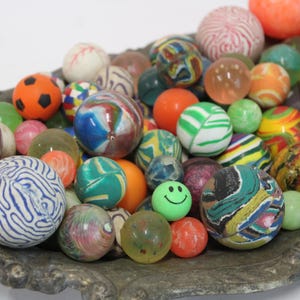 May include: A collection of numerous multi-colored rubber bouncy balls in various sizes and patterns, including solid colors, swirls, and a green ball with a smiley face, all resting together in an ornate, weathered, metallic oval-shaped tray.