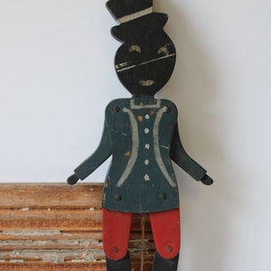 May include: A vintage articulated wooden jumping jack toy featuring a figure in a black top hat, a dark blue jacket with white painted details, red breeches, and black legs. The figure has metal fasteners at the joints and a pull string.