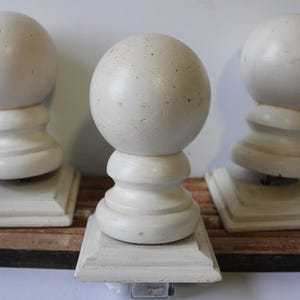 May include: Three off-white, decorative wooden finials with a ball top and a square base. The finials are mounted on a rectangular, stained wooden base. The design is simple and elegant, suitable for home decor.