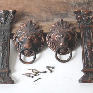 May include: Bronze-colored decorative hardware set featuring two lion head door knockers with ring handles and two column-shaped supports. Includes screws and nails for installation. The hardware has an antique finish.