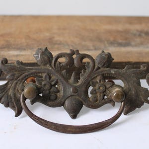 May include: Antique bronze drawer pull with ornate floral and leaf design. The pull features a curved handle and two small knobs. The hardware is mounted on a white surface with a wooden background.