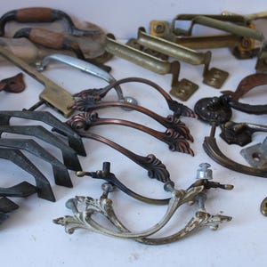 May include: A collection of assorted metal drawer pulls and cabinet handles in various finishes including brass, copper, silver, and dark iron. The hardware features diverse shapes, ranging from simple straight bars to ornate, curved, and decorative designs.