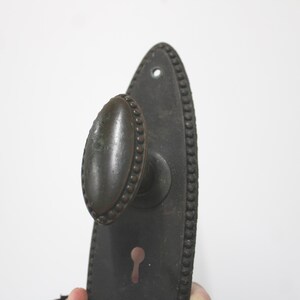 May include: A dark brown metal door handle set featuring an oval-shaped knob and a matching elongated backplate. The backplate has a pointed top and bottom, a beaded border, and a keyhole cutout. The knob also features a beaded edge detail.