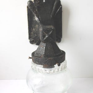 May include: A wall-mounted light fixture featuring a black, textured cast iron base with a rectangular backplate and a clear, bulbous glass shade. The glass shade has a beaded texture around the top and bottom rims, secured by small metal screws.