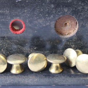 May include: A collection of brass mushroom-shaped knobs with a shiny finish. The knobs are arranged in a row, with a dark background. There are also two holes, one red and one brown, in the background.