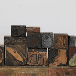 May include: A collection of rectangular wooden letterpress blocks featuring various engraved designs including a feather, a boot, a violin, an open book, a circular portrait, and text such as REALTORS, arranged in a stacked formation.